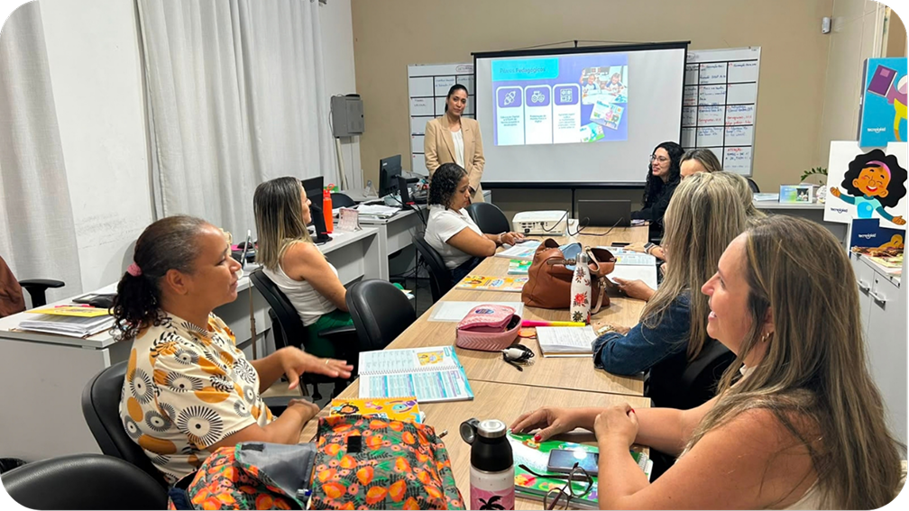 Formadora em pé diante de uma projeção conduz encontro com professoras sentadas em volta de uma mesa com materiais didáticos