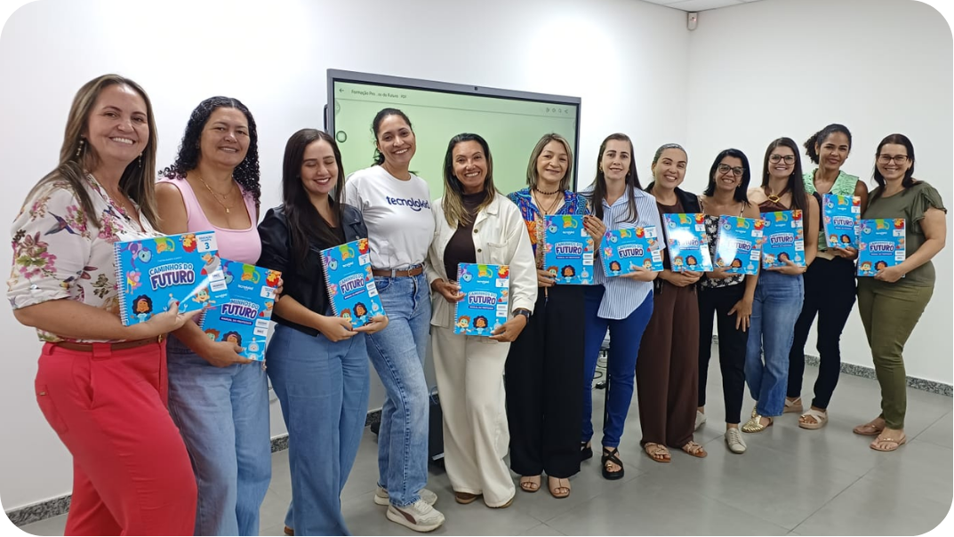 Professoras em pé ao lado de uma tela interativa segurando livros da coleção Caminhos do Futuro